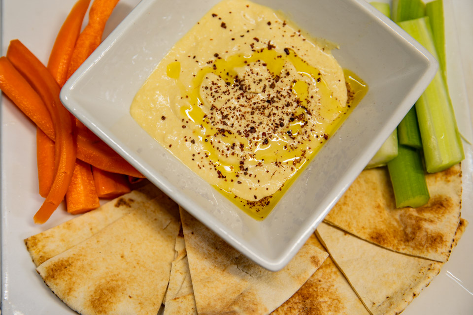 A plate of creamy hummus served at Simone on Sunset in Rice Village, Houston, accompanied by fresh pita bread, celery sticks, and carrot sticks.