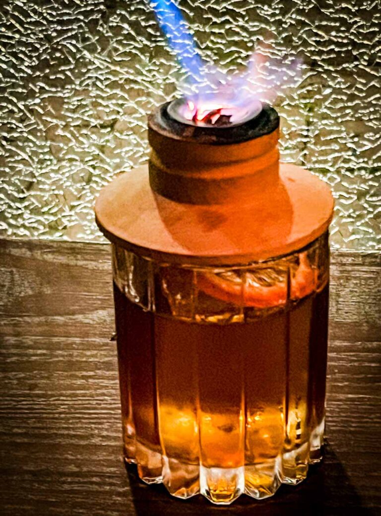 A glass of Black Walnut Old Fashioned cocktail at Simone on Sunset, featuring Angel's Envy Bourbon, black walnut liqueur, turbinado sugar, and Angostura and mole bitters, garnished with a hint of smoked cinnamon.